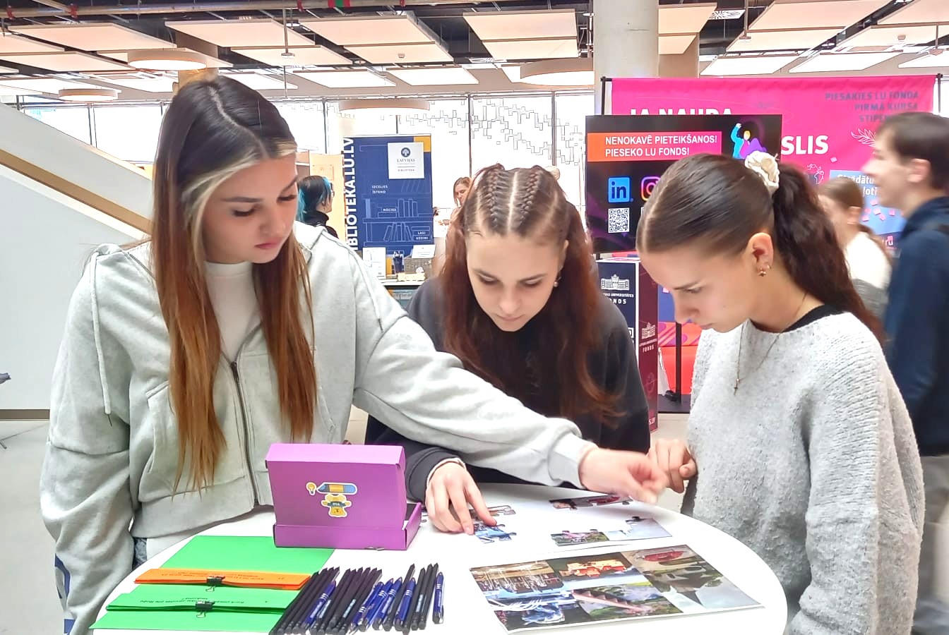 Meitenes pie reģionālo filiāļu stenda kopīgi saliek filiāļu pilsētu puzli