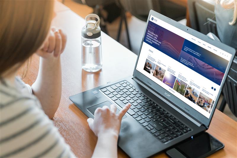 Cilvēks sēž pie galda un lieto portatīvo datoru, kura ekrānā atvērta zinātnes portāla tīmekļvietne ar rakstu virsrakstiem un attēliem. Uz galda redzama ūdens pudele, apkārt – auditorijas vai mācību telpas krēsli.