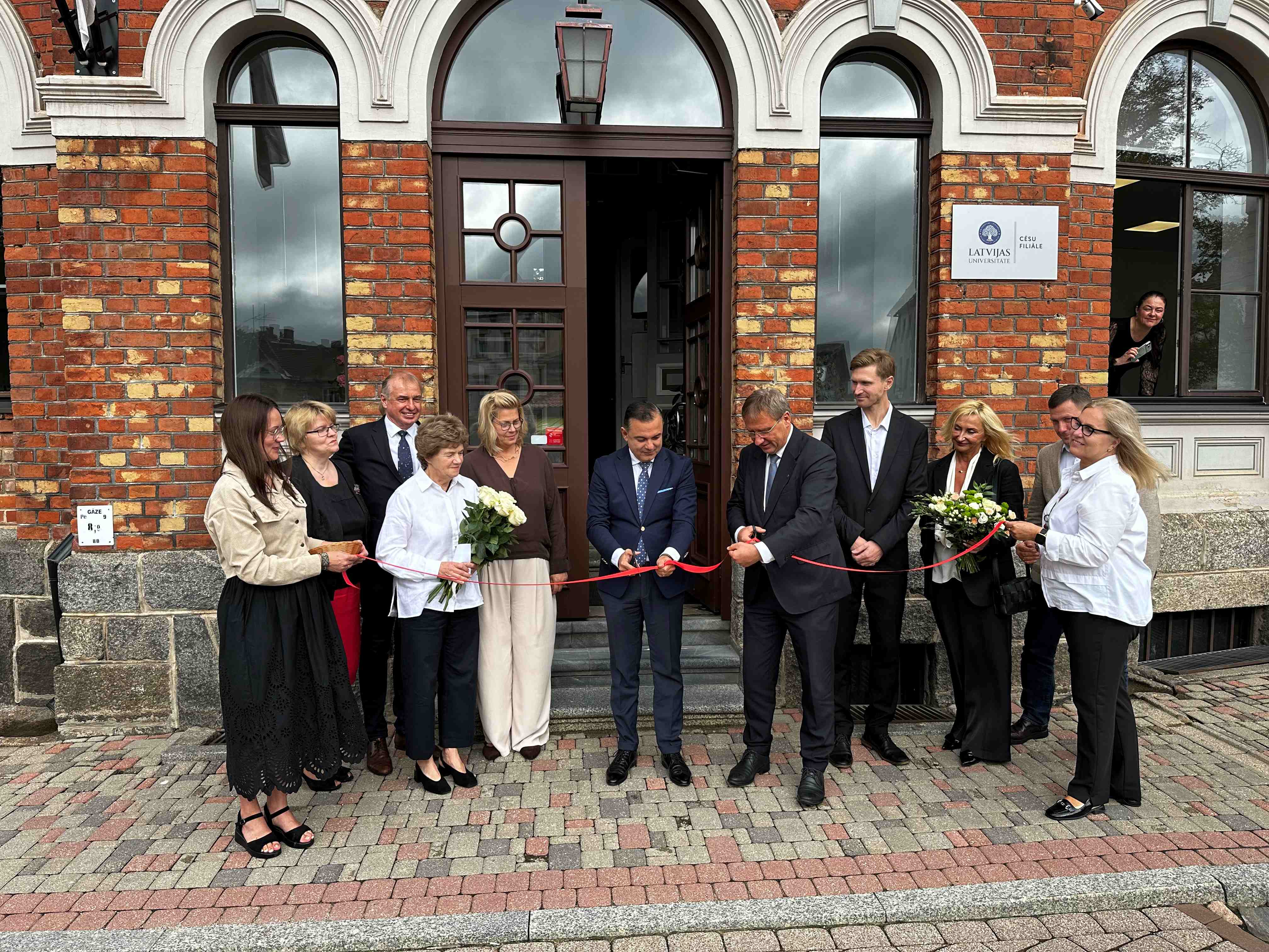 Pie Latvijas Universitātes Cēsu filiāles ēkas ieejas notiek simboliska lentes griešanas ceremonija, kurā piedalās vairāki cilvēki. Virs ieejas redzama norāde uz Latvijas Universitātes Cēsu filiāli.